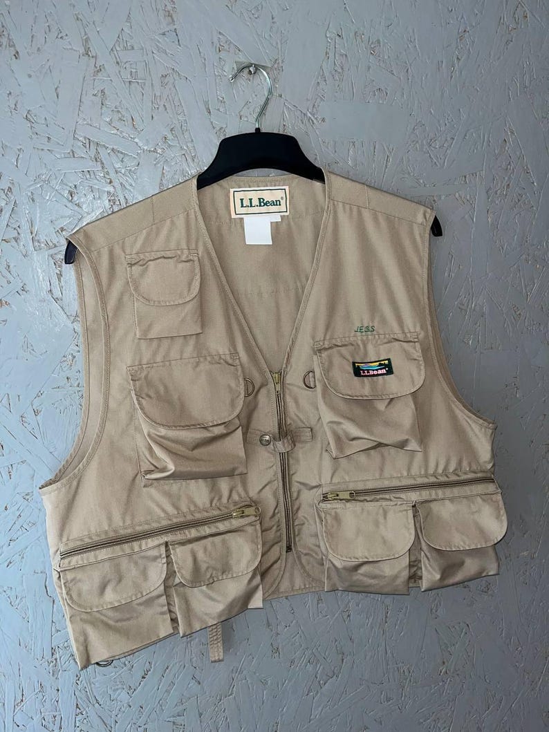 May include: A tan utility vest featuring multiple pockets, a front zipper closure, and an L.L.Bean brand label at the neckline. The garment includes a small embroidered name tag reading JESS and a rectangular L.L.Bean logo patch on the chest pocket.