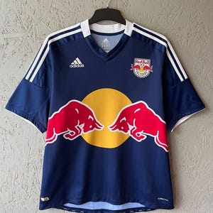Peut inclure: Un maillot de football Adidas bleu marine avec des rayures blanches sur les épaules et un col en V. Il présente le logo de l'équipe Red Bull Salzbourg et un cercle jaune avec deux silhouettes de taureaux rouges.
