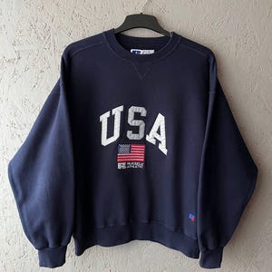 以下が含まれることがあります： ネイビーブルーのスウェットシャツ。白いブロック体で「USA」の文字と小さなアメリカ国旗のグラフィックがプリントされています。「Russell Athletic」のロゴも表示されています。