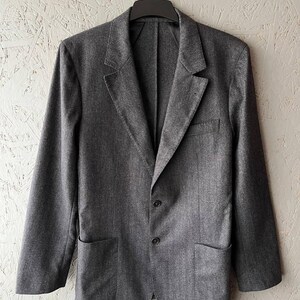 Puede incluir: Un blazer gris con dos bolsillos delanteros y dos botones, colgado de una percha negra. El blazer tiene solapas con muescas y un corte entallado. La tela parece ser un material tejido.