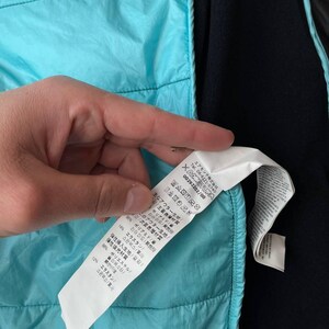May include: A close-up of a turquoise quilted jacket with a black interior. A white tag with Japanese text and care symbols is visible. The jacket is made of polyamide and elastane.