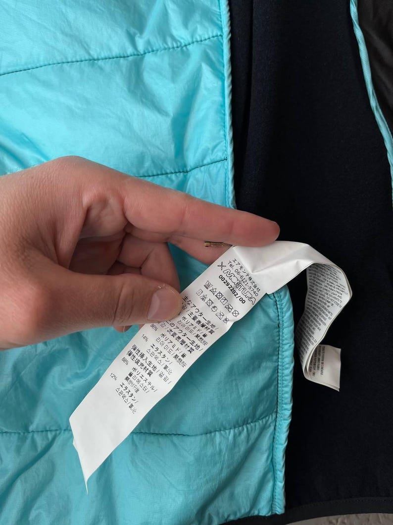 May include: A close-up of a turquoise quilted jacket with a black interior. A white tag with Japanese text and care symbols is visible. The jacket is made of polyamide and elastane.