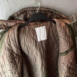K&ouml;nnte beinhalten: Eine olivgr&uuml;ne Jacke mit gestepptem braunem Innenfutter. Das Kleidungsst&uuml;ck verf&uuml;gt &uuml;ber eine Kapuze, einen schwarzen Kunststoffkleiderb&uuml;gel und ein leeres, wei&szlig;es, rechteckiges Etikett in der Mitte des inneren R&uuml;ckenteils. Mit Metalldruckkn&ouml;pfen.