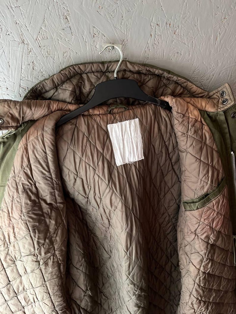 K&ouml;nnte beinhalten: Eine olivgr&uuml;ne Jacke mit gestepptem braunem Innenfutter. Das Kleidungsst&uuml;ck verf&uuml;gt &uuml;ber eine Kapuze, einen schwarzen Kunststoffkleiderb&uuml;gel und ein leeres, wei&szlig;es, rechteckiges Etikett in der Mitte des inneren R&uuml;ckenteils. Mit Metalldruckkn&ouml;pfen.
