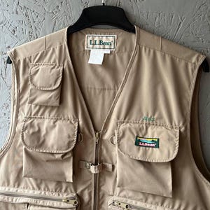 May include: A tan L.L.Bean utility vest featuring multiple front pockets, a central zipper closure, and a small embroidered logo patch on the chest pocket. The garment includes a collar tag and a small white care label hanging from the neckline.