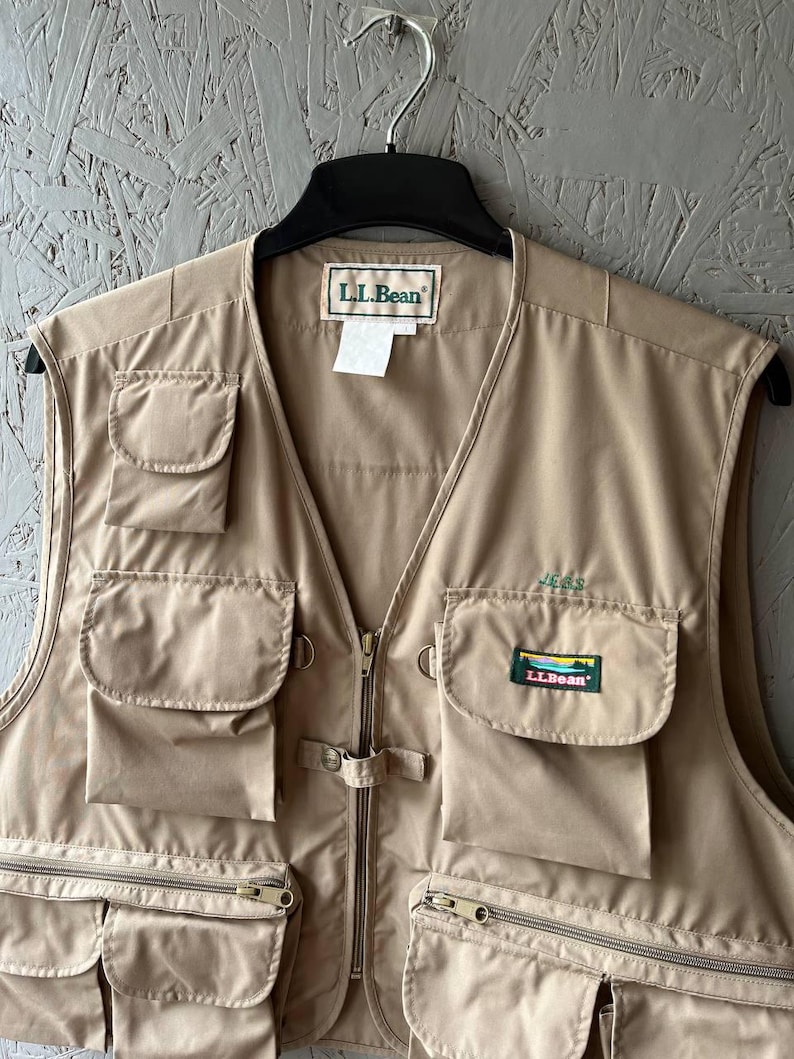 May include: A tan L.L.Bean utility vest featuring multiple front pockets, a central zipper closure, and a small embroidered logo patch on the chest pocket. The garment includes a collar tag and a small white care label hanging from the neckline.