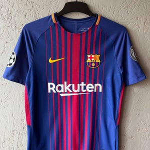 Puede incluir: Una camiseta de fútbol a rayas azules y rojas con la palabra "Rakuten" en blanco en la parte delantera. La camiseta tiene un logotipo de Nike amarillo y un escudo del equipo. Las mangas tienen un parche de la Liga de Campeones.