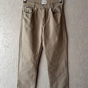 May include: Tan-colored denim jeans with a straight leg fit, hanging on a metal hanger. The jeans feature classic five-pocket styling, a button and zipper closure, and the D&G logo on the back pocket.