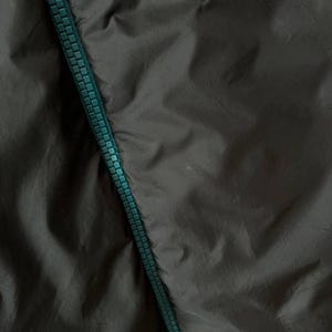 May include: Close-up of dark grey fabric with a teal zipper running diagonally. The fabric has a slightly wrinkled texture, and the zipper teeth are clearly visible.