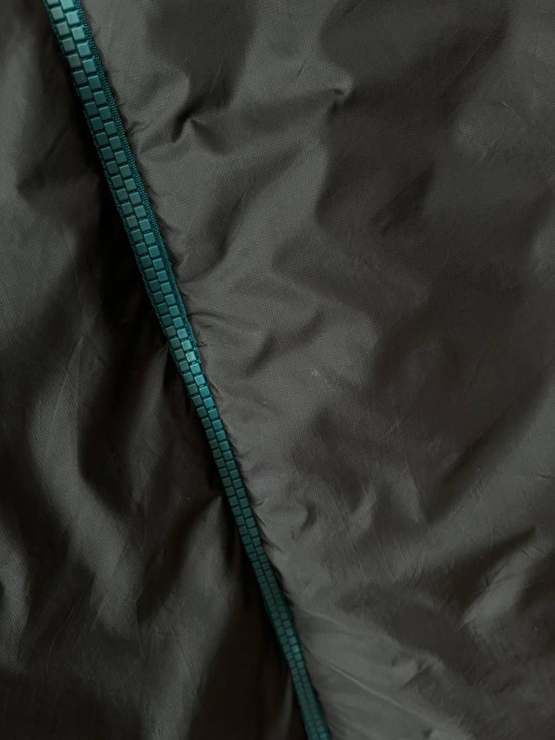 May include: Close-up of dark grey fabric with a teal zipper running diagonally. The fabric has a slightly wrinkled texture, and the zipper teeth are clearly visible.