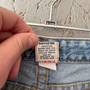 Puede incluir: Primer plano de la cintura de unos vaqueros azules. Una etiqueta blanca con el texto "RN#112790" e instrucciones de cuidado es visible. Los vaqueros est&aacute;n colgados de una percha met&aacute;lica.