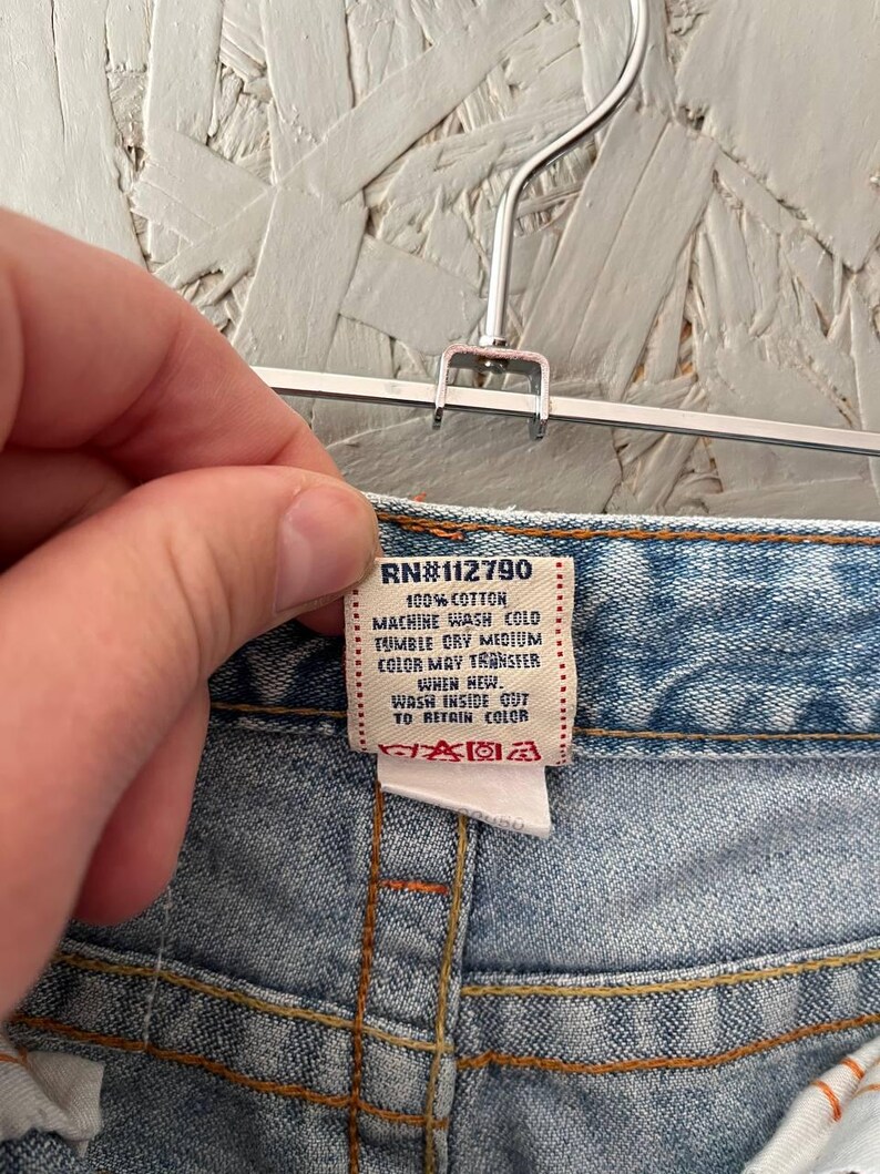 Puede incluir: Primer plano de la cintura de unos vaqueros azules. Una etiqueta blanca con el texto "RN#112790" e instrucciones de cuidado es visible. Los vaqueros est&aacute;n colgados de una percha met&aacute;lica.