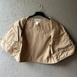 May include: A tan utility vest featuring multiple front pockets, a zippered compartment on the left side, and a rectangular L.L. Bean brand label sewn into the inner neckline. The vest is sleeveless and constructed from a smooth, solid-colored fabric.