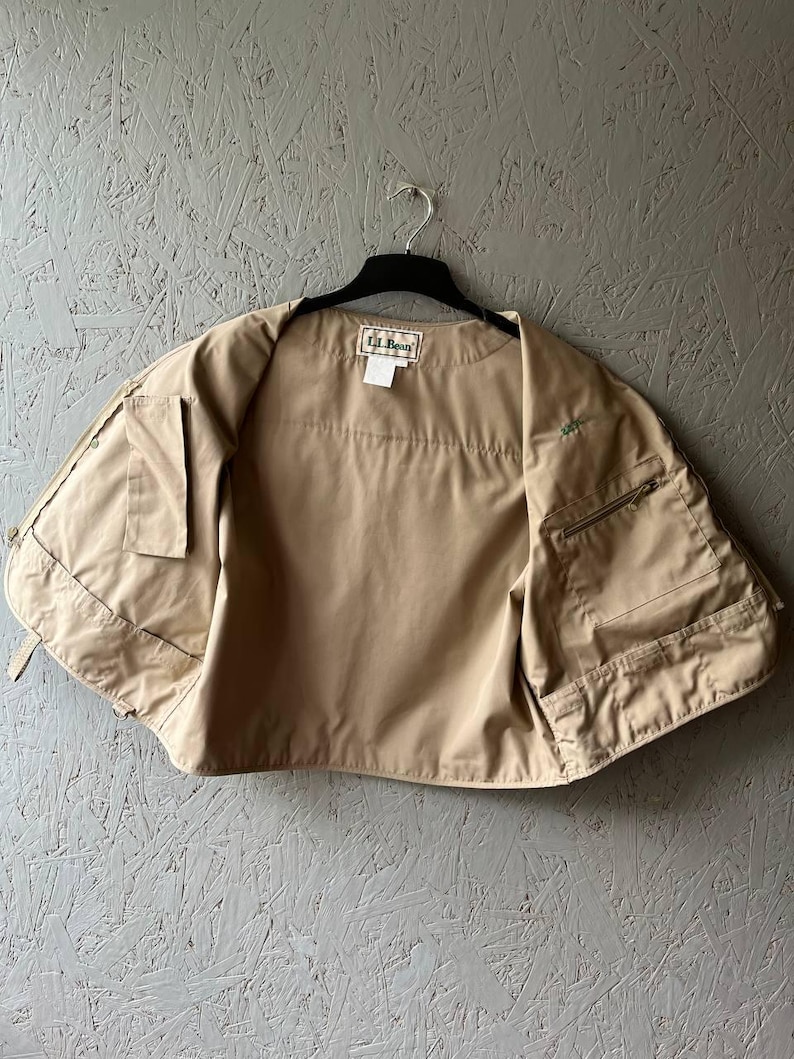 May include: A tan utility vest featuring multiple front pockets, a zippered compartment on the left side, and a rectangular L.L. Bean brand label sewn into the inner neckline. The vest is sleeveless and constructed from a smooth, solid-colored fabric.