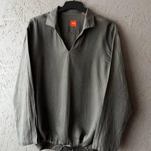 May include: A gray long-sleeve shirt with a V-neck collar and a drawstring at the waist. The shirt has a small orange tag with the brand name "BOSS" on the chest. The shirt is hanging on a black hanger.
