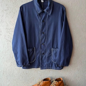 Puede incluir: Una chaqueta de trabajo azul marino con costuras blancas y botones negros cuelga sobre un par de botas de cuero marrón con suelas blancas. La chaqueta tiene dos bolsillos delanteros y un cuello. Las botas están atadas y colocadas sobre una superficie de madera.