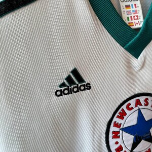 K&ouml;nnte beinhalten: Wei&szlig;es Adidas-Fu&szlig;balltrikot mit schwarzen und gr&uuml;nen Akzenten. Mit Adidas-Logo und Newcastle Brown Ale-Abzeichen. Das Trikot hat einen V-Ausschnitt und schwarze Streifen auf den Schultern.