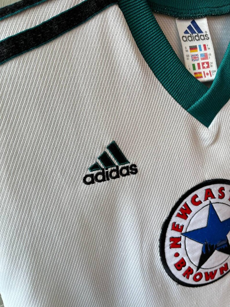 K&ouml;nnte beinhalten: Wei&szlig;es Adidas-Fu&szlig;balltrikot mit schwarzen und gr&uuml;nen Akzenten. Mit Adidas-Logo und Newcastle Brown Ale-Abzeichen. Das Trikot hat einen V-Ausschnitt und schwarze Streifen auf den Schultern.