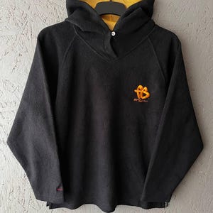 以下が含まれることがあります： イエローの裏地が付いたフードとネックラインにボタン留めが付いた黒いフリースパーカー。 パーカーの胸には、オレンジと黄色の小さなロゴが付いています。 長袖トップスはリラックスしたフィット感です。