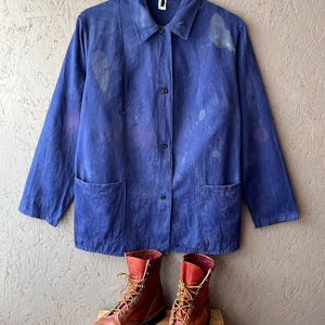 May include: A blue work jacket with a collar, long sleeves, four front buttons, and two large patch pockets, featuring faded areas and paint splatters. Below the jacket are a pair of brown leather lace-up work boots with yellow laces.