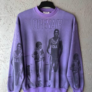 May include: A purple crew neck sweatshirt with the word "OPENAIR" printed across the chest in black. The sweatshirt features a graphic of people running and walking. The sleeves also have the word "OPENAIR" printed on them.