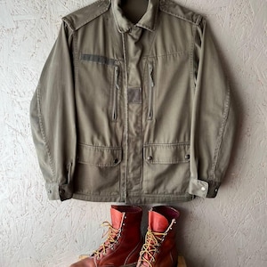 May include: An olive green utility jacket with a collared neckline, shoulder epaulets, two vertical zippered chest pockets, and two lower button-flap pockets, paired with a pair of brown leather lace-up work boots with yellow and red laces.
