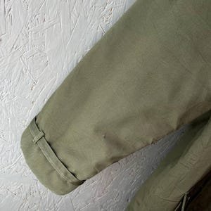 K&ouml;nnte beinhalten: Ein olivgr&uuml;ner Jacken&auml;rmel mit einem strukturierten &Auml;rmelabschluss, einem passenden Stoffriegel und einem kleinen Knopfdetail. Der Stoff hat eine glatte, matte Oberfl&auml;che und eine sichtbare Naht entlang des &Auml;rmels, was ein schlichtes Design zeigt.