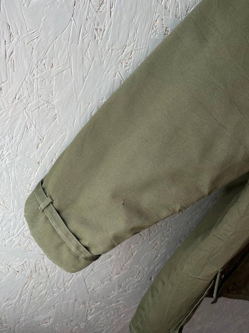 K&ouml;nnte beinhalten: Ein olivgr&uuml;ner Jacken&auml;rmel mit einem strukturierten &Auml;rmelabschluss, einem passenden Stoffriegel und einem kleinen Knopfdetail. Der Stoff hat eine glatte, matte Oberfl&auml;che und eine sichtbare Naht entlang des &Auml;rmels, was ein schlichtes Design zeigt.