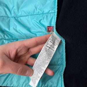 May include: Close-up of a turquoise quilted jacket lining and black fleece material. A small tag with the word "PRIMALOFT" is visible. A white tag with care instructions is also visible.