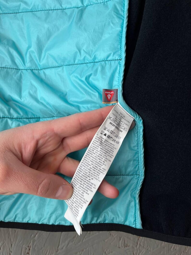 May include: Close-up of a turquoise quilted jacket lining and black fleece material. A small tag with the word "PRIMALOFT" is visible. A white tag with care instructions is also visible.