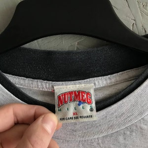 Puede incluir: Una camiseta gris con cuello de canal&eacute; negro. La camiseta tiene una etiqueta blanca con el texto "NUTMEG MILLS" y "XL" en rojo. La etiqueta tambi&eacute;n dice "FOR CARE SEE REVERSE" en negro.