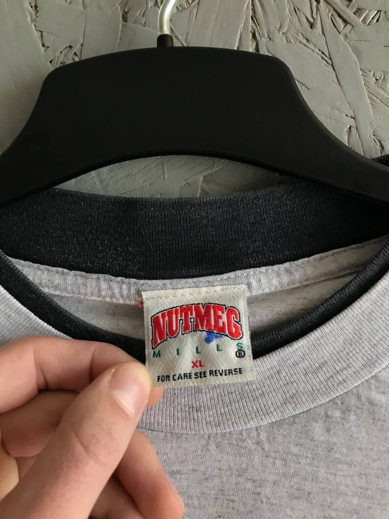 Puede incluir: Una camiseta gris con cuello de canal&eacute; negro. La camiseta tiene una etiqueta blanca con el texto "NUTMEG MILLS" y "XL" en rojo. La etiqueta tambi&eacute;n dice "FOR CARE SEE REVERSE" en negro.