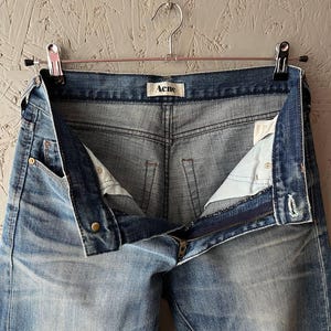 以下が含まれることがあります： 部分的にジッパーが開いた、金属製のハンガーに吊るされたライトブルーのデニムジーンズ。ジーンズは色あせたウォッシュ加工で、ブランド名「Acne」がウエストバンドに表示されています。