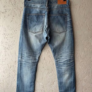 以下が含まれることがあります： 色褪せたウォッシュ加工が施されたライトブルーデニムのジーンズ。革製のパッチと、使用感のある跡が見えます。金属製のハンガーに吊るされており、バックポケットと縫い目がはっきりと見えます。