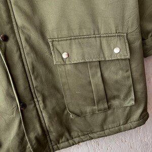 K&ouml;nnte beinhalten: Eine olivgr&uuml;ne Jacke mit einer gro&szlig;en aufgesetzten Vordertasche mit Klappe und zwei silberfarbenen Metalldruckkn&ouml;pfen. Die Jacke verf&uuml;gt &uuml;ber eine vertikale Knopfleiste an der Seite mit dunkelbraunen Druckkn&ouml;pfen und sichtbaren N&auml;hten.