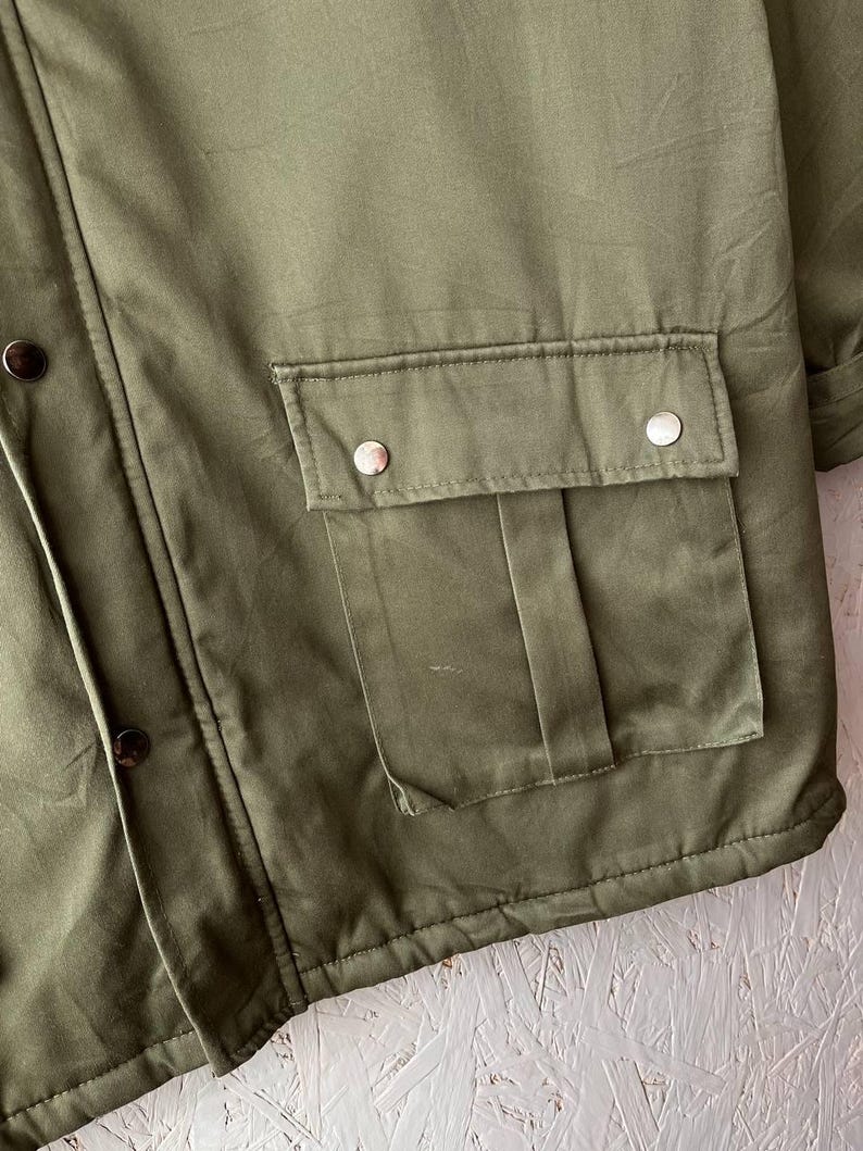 K&ouml;nnte beinhalten: Eine olivgr&uuml;ne Jacke mit einer gro&szlig;en aufgesetzten Vordertasche mit Klappe und zwei silberfarbenen Metalldruckkn&ouml;pfen. Die Jacke verf&uuml;gt &uuml;ber eine vertikale Knopfleiste an der Seite mit dunkelbraunen Druckkn&ouml;pfen und sichtbaren N&auml;hten.