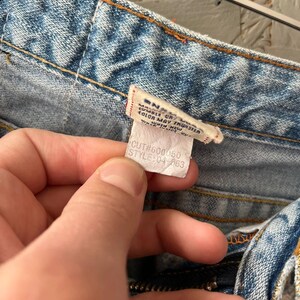 Puede incluir: Primer plano de unos vaqueros de mezclilla azul claro con una etiqueta. La etiqueta dice "MANUFACTURED BY... COLOR MAY TRANSFER... WESTERN NEW YORK... CUT#600050 STYLE:04-563". Los vaqueros tienen cremallera y costuras naranjas.