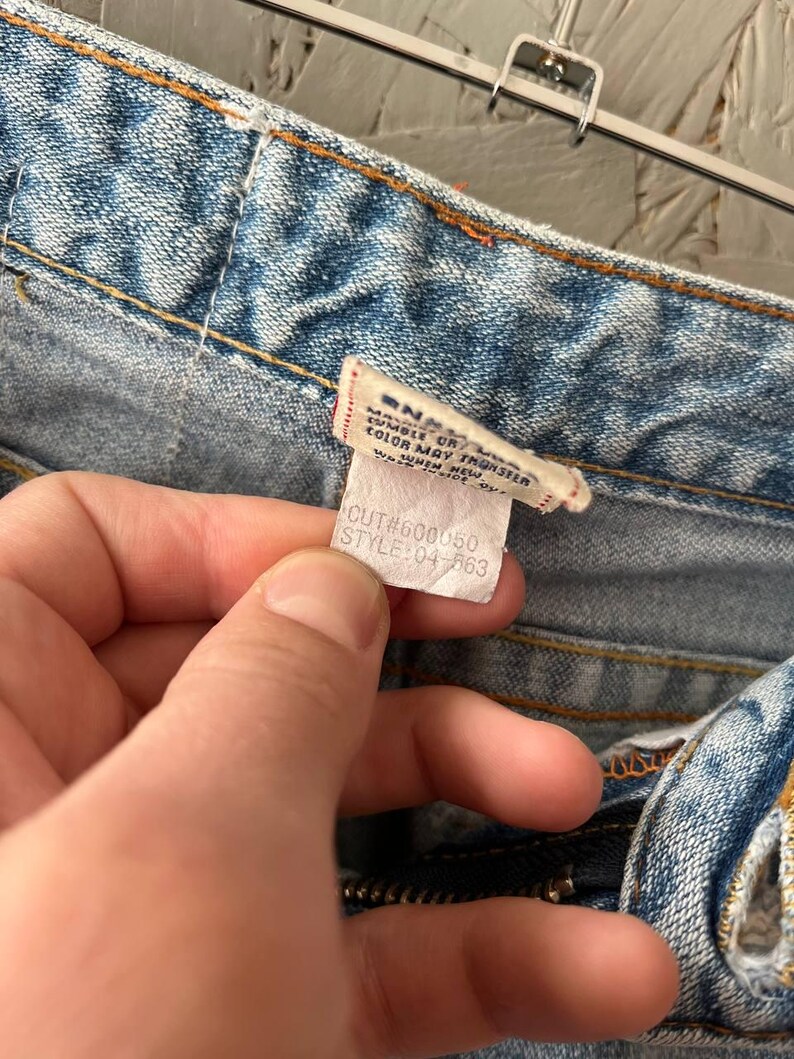 Puede incluir: Primer plano de unos vaqueros de mezclilla azul claro con una etiqueta. La etiqueta dice "MANUFACTURED BY... COLOR MAY TRANSFER... WESTERN NEW YORK... CUT#600050 STYLE:04-563". Los vaqueros tienen cremallera y costuras naranjas.