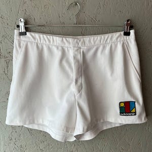 May include: White athletic shorts with a small embroidered logo on the right leg. The logo features a square with a yellow circle, red triangle, blue rectangle, and green stripe. The shorts have a button closure and pockets.