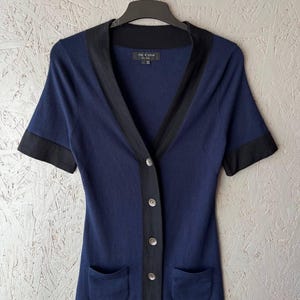 Puede incluir: Un cárdigan azul marino de manga corta con ribetes y botones negros. El cárdigan tiene cuello en V, dos bolsillos delanteros y está colgado de una percha negra. El nombre de la marca es visible en la etiqueta.