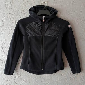 Puede incluir: Chaqueta de forro polar negra con capucha y cremallera completa. La chaqueta presenta un panel de nailon acolchado en el pecho y un pequeño parche con el logotipo en la manga superior. Colgada en una percha negra.