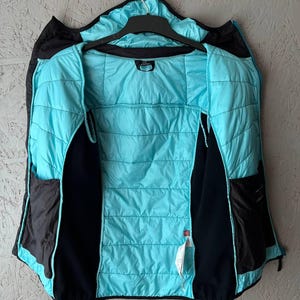 May include: A black and turquoise quilted jacket hanging on a black hanger. The jacket has a turquoise interior and black exterior with two black pockets. The jacket is open, revealing the interior lining.