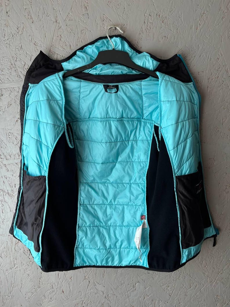 May include: A black and turquoise quilted jacket hanging on a black hanger. The jacket has a turquoise interior and black exterior with two black pockets. The jacket is open, revealing the interior lining.