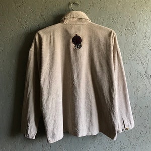 May include: A beige corduroy shirt with a long sleeve and a button-up front. The shirt has a small embroidered patch on the back with a skull and crossbones design.