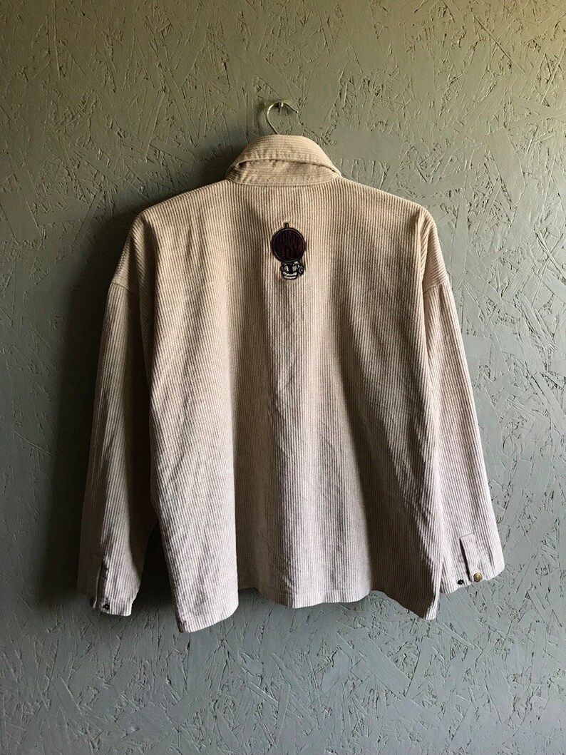 May include: A beige corduroy shirt with a long sleeve and a button-up front. The shirt has a small embroidered patch on the back with a skull and crossbones design.
