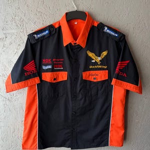 May include: Black and orange short-sleeve button-up shirt with embroidered logos. Features "GoldWing" and "Honda" branding, along with "Michelin" and other sponsor patches. The shirt has a classic workwear style with two chest pockets.