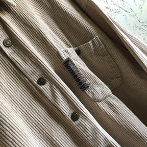 May include: A beige corduroy shirt with a patch pocket on the left side. The pocket has a colorful "homieboy" label.