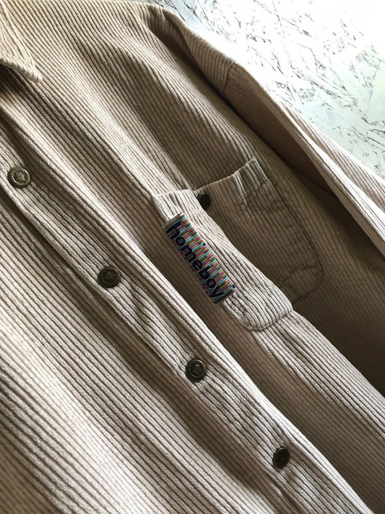 May include: A beige corduroy shirt with a patch pocket on the left side. The pocket has a colorful "homieboy" label.