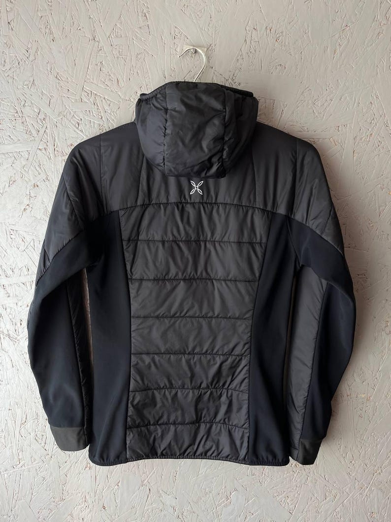 May include: Black hooded jacket with a quilted central panel and solid black side panels. The jacket has a zip closure and a small white logo on the upper back. The jacket is hanging on a silver hanger.