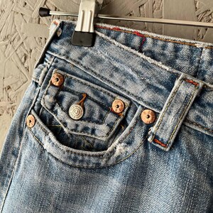 Puede incluir: Primer plano de unos vaqueros de mezclilla azul claro con remaches de color cobre y un peque&ntilde;o bolsillo delantero. Los vaqueros est&aacute;n desgastados, mostrando signos de uso. Las costuras son de color naranja en contraste. Los vaqueros cuelgan de una percha met&aacute;lica.